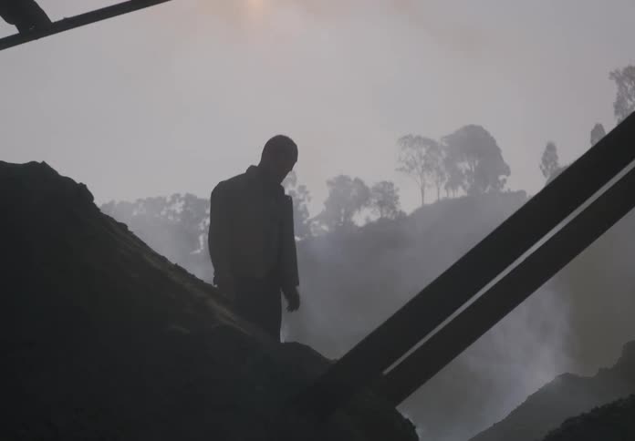 Man is silhouetted by dust on sunny day as he walks across mounds of gravel and dirt at plant