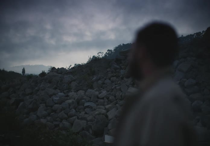 A man stands in front of a mountain of large rocks while staring at the gray cloudy sky