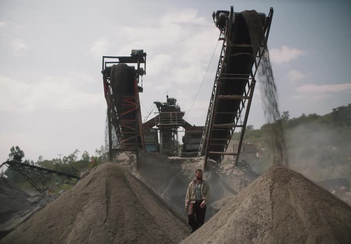 A man stands by piles of dirt under conveyor belts and looks around