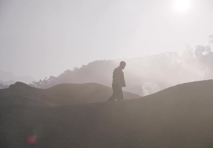 A man walks across stone piles as a machine is working and the sun shines on smoke moving across the area