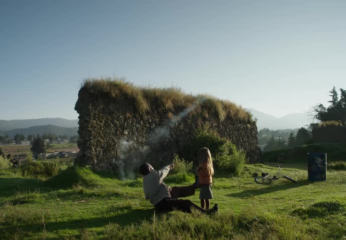 A man is outside sitting in the grass with his young daughter standing next to him near ruins of a wall as he lights a firecracker and throws it and she runs to the smoke where it exploded