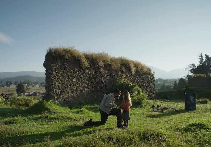 A young girl stands by her dad as he kneels in a grassland and they laugh together