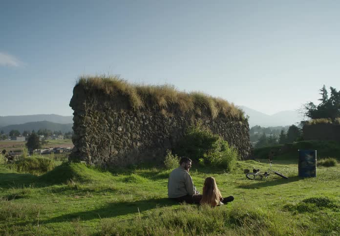 A man is outside with his young daughter near ruins of a wall and he sits down and lights a firecracker and throws it