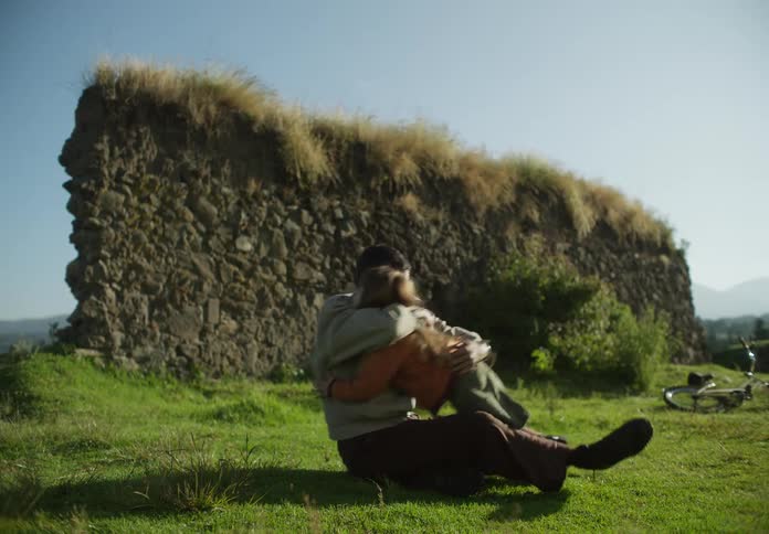 Man lights cigarette and throws it away then hugs daughter as they sit on ground with mountains in the background