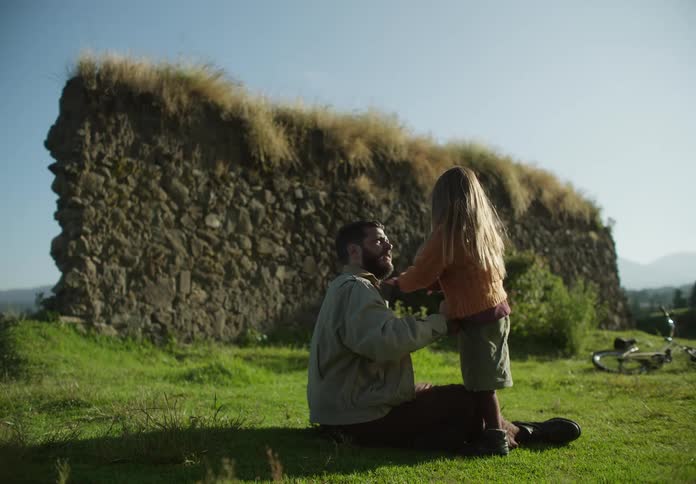 Father and daughter sitting on green grass near an abandoned rock wall playing at morning