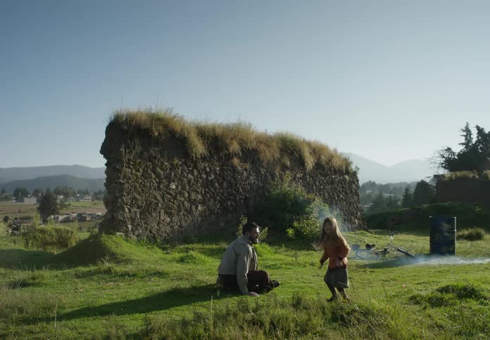 A man is outside sitting in the grass with his young daughter standing next to him near ruins of a wall as he lights a firecracker and throws it and she walks to the smoke where it exploded