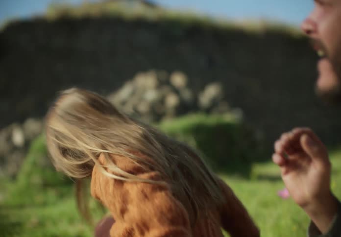 A man is sitting outside in the grass with his young daughter as he pulls a firecracker out of his pocket and shows her then he lights it and throws it and she stands up and he hugs her