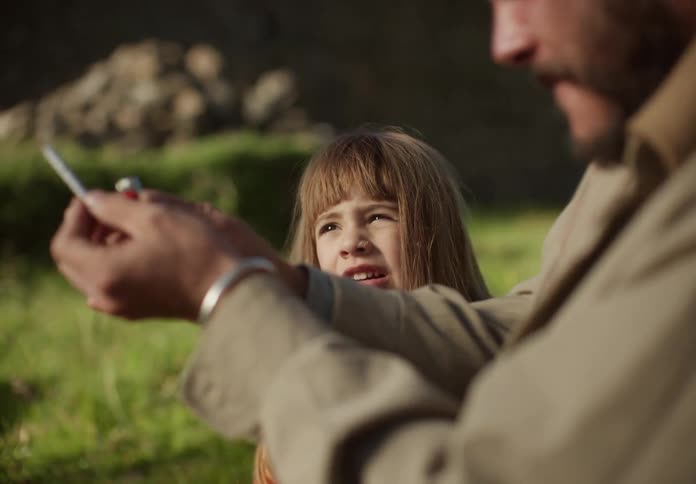 A young girl sits in a grassy field and talks with her father while he lights a firecracker and throws it