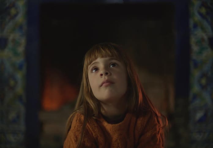Redheaded little girl looks around while sitting with her back to the lit fireplace