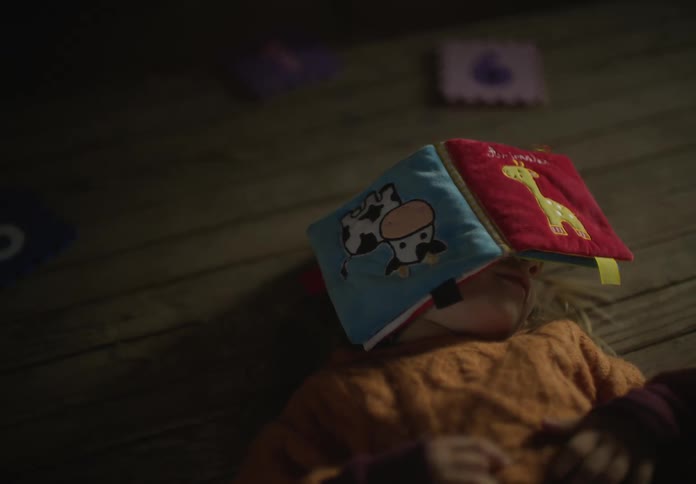 A child lies on the floor with a soft book on her face and laughs as her dad touches her hand