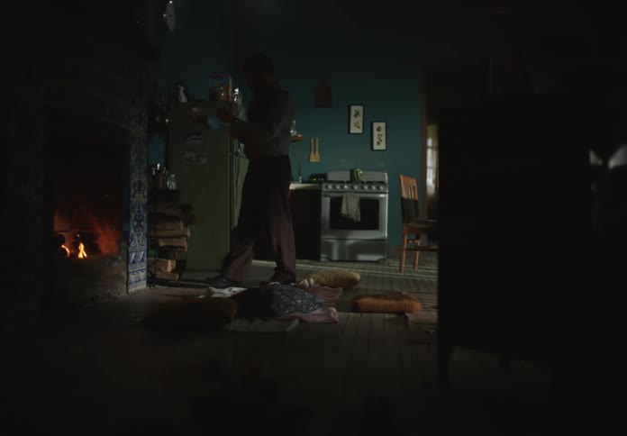 A man takes off his jacket as he enters the house then rolls up his sleeves and walks to the fireplace to poke at the fire