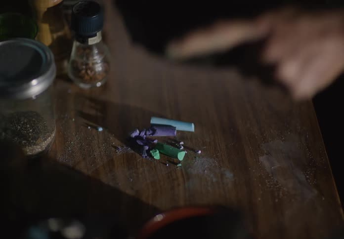 A person uses an iron skillet to crush pieces of colored chalk on a table