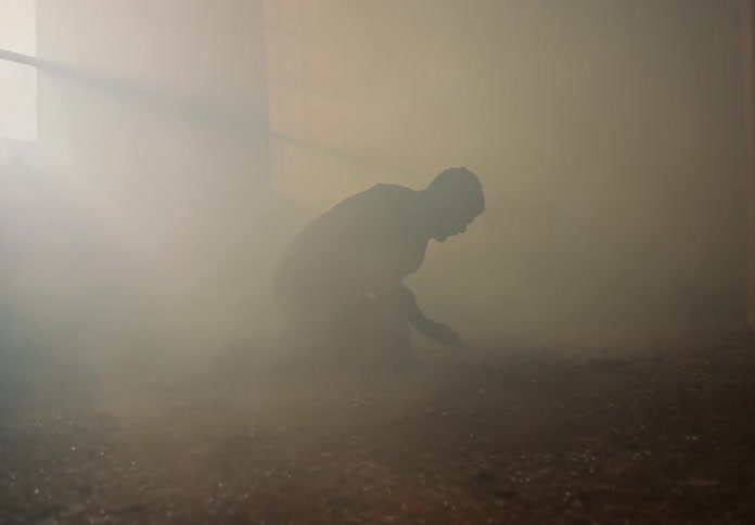 Man laying on the floor of a room full of smoke shakily stands up
