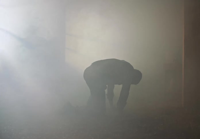A man gets up from the ground and stumbles around a smoke filled room