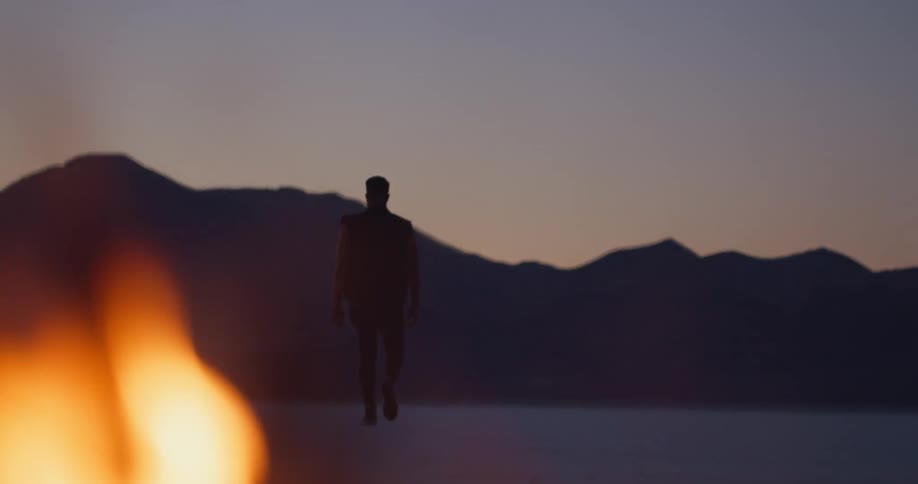 A man looking at a campfire turns and walks away towards the mountains