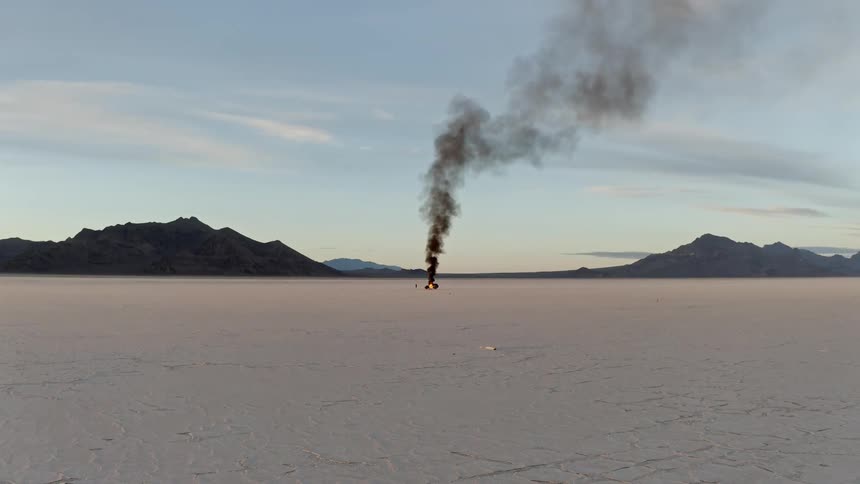 Black smoke rises from an automobile on fire in the desert near mountains as a man stands watching the flames burn