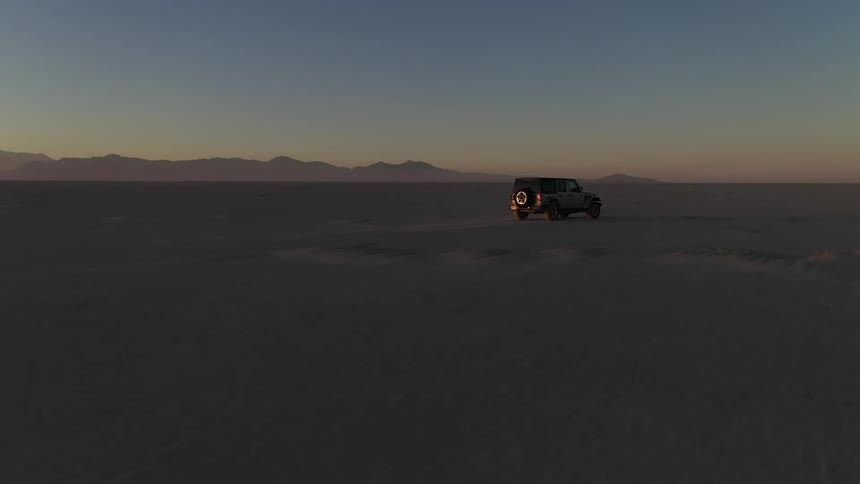 A jeep is traveling across the desert and there are mountains in the distance under a hazy sky