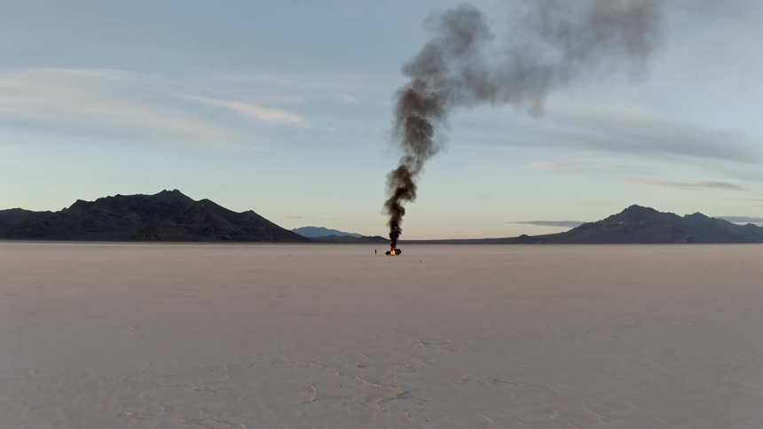 Black smoke rises from an automobile on fire in the desert as a man stands watching the flames burn