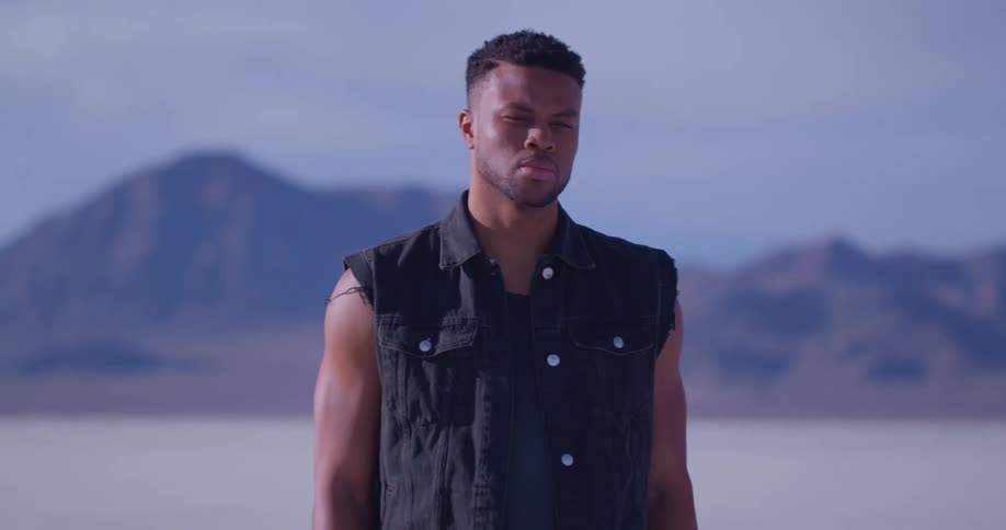 A man stands in front of a mountain range and looks off in to the distance