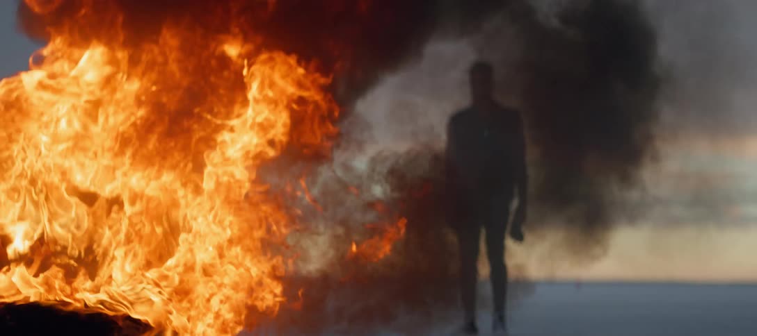 Silhouette of a man standing in the plains of a desert in front of a burning tire