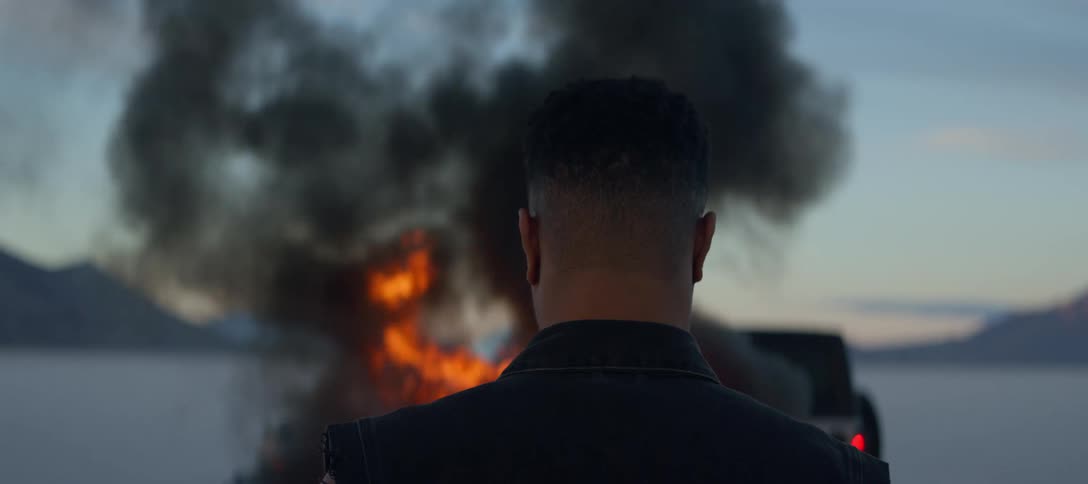 Man stands in front of fire near car parked on plain stretching to mountains under red horizon