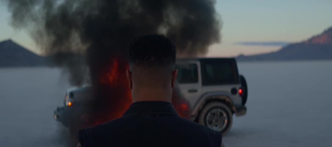 A man watches motionlessly as a nearby vehicle is consumed by fire in a desert in the evening