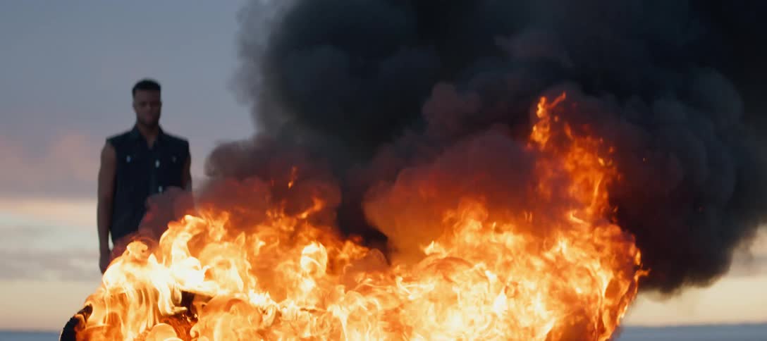 A man watches as flames consume an object while standing outdoors in the evening