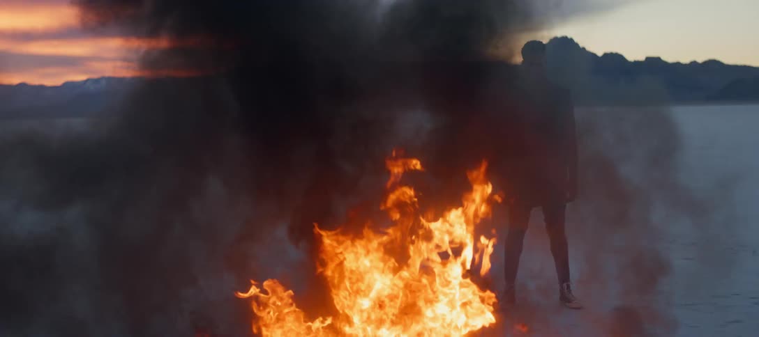 Man stands in front of mountain range looking at bonfire billowing smoke