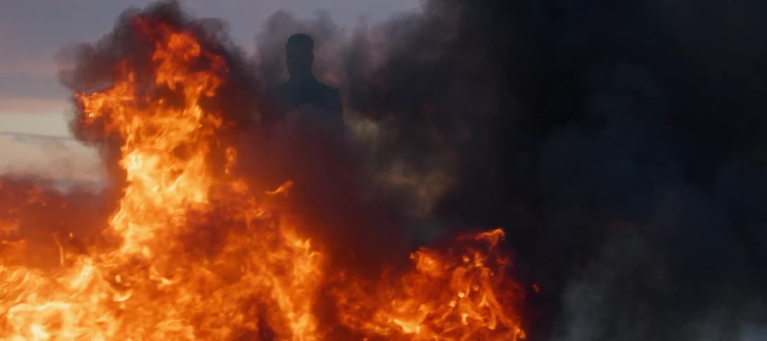 A man is standing outside watching a blazing fire as black smoke rises from the flames
