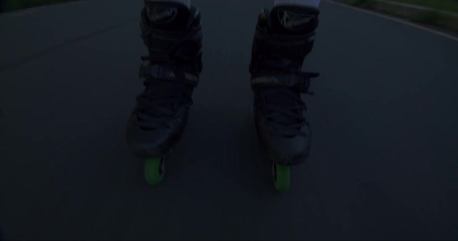 A man wearing roller blades with green wheels is rolling down a street and they go through a dark area