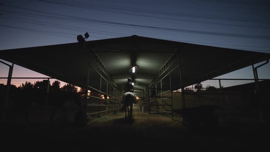 A woman rides a horse into a barn with stalls