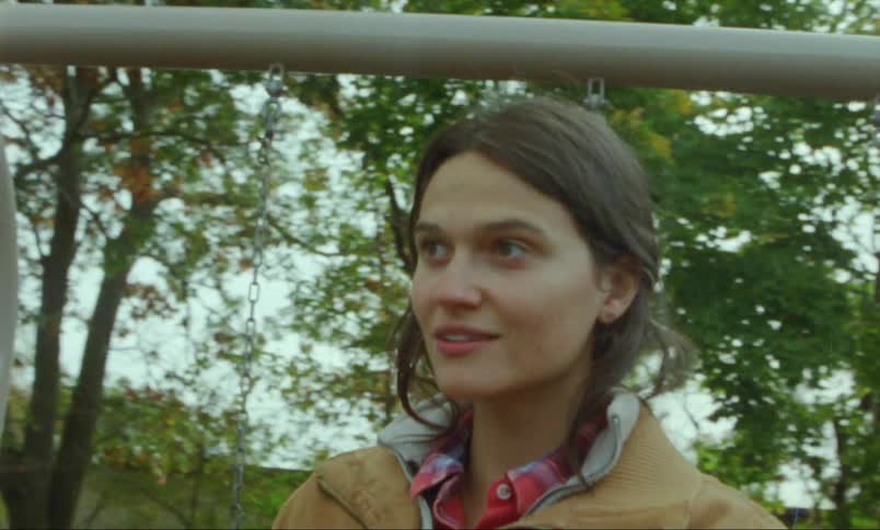 A woman is standing outside at a playground near swings as she speaks to someone with a smile on her face