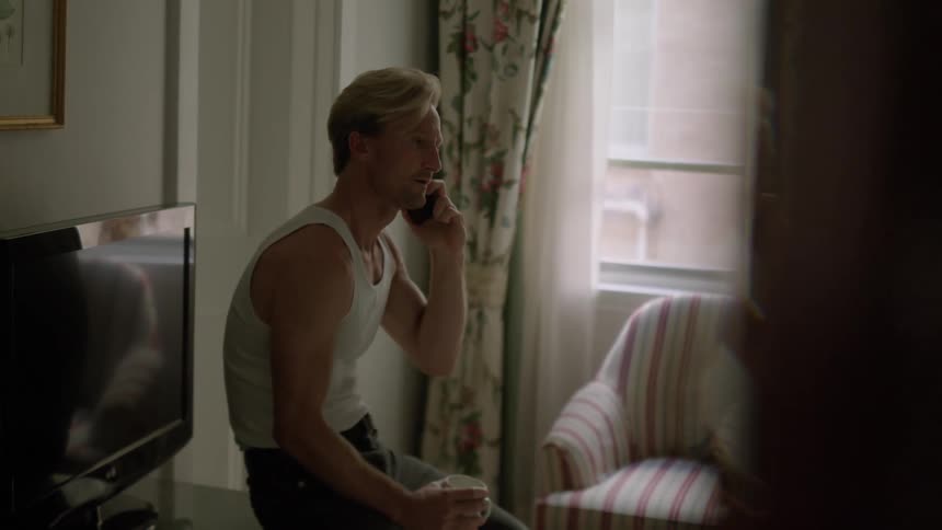 Blonde man talking on smartphone in living room sits down on television stand