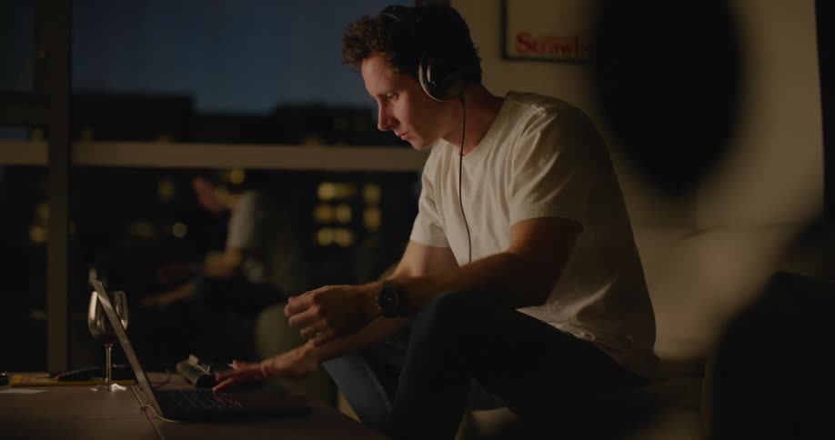 A man sitting on his couch puts on headphones and leans forward to look at his laptop screen