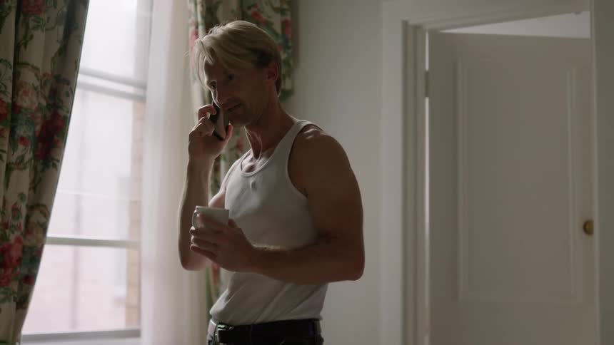 A man talking on the phone and drinking a cup of coffee next to a window with curtains at home