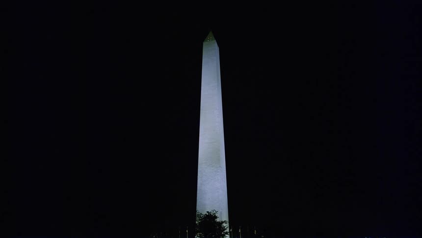 Trees are silhouetted against highlighted washington monument at night