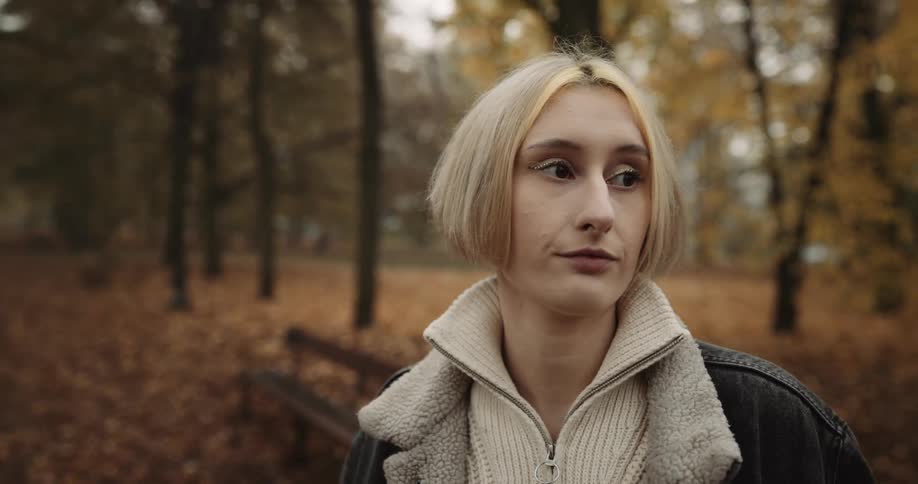 Blonde woman with bob haircut white sweater denim jacket and jeans waits for her partner in public park near the benches