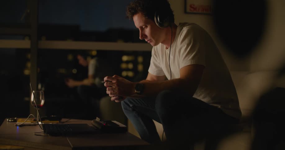 A man wearing headphones is sitting at a table with a laptop computer and playing a small electronic keyboard as he picks up his phone and scrolls with his thumbs