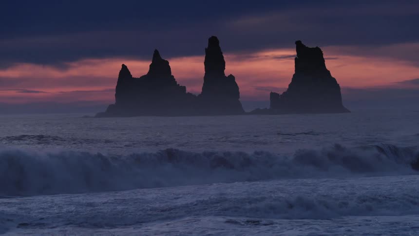 Iceland Ocean waves roll into a beach as a large rock formation looms off shore at sunset