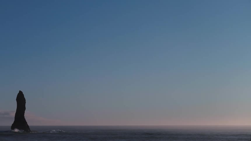 A rock formation stands tall in the water as wave roll in under a clear hazy sky