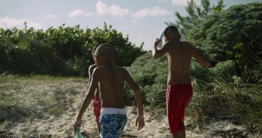 A group of boys in swimming trunk walk up a sandy hill together during the day