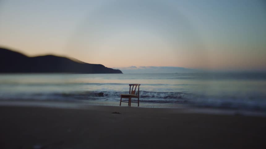 A chair is sitting on a beach as waves roll into shore and under its legs in the sand