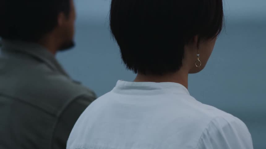 Woman with short hair earrings and white blouse sits next to man in green shirt watching sea