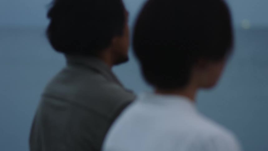 Man in olive drab shirt and woman wearing white shirt sit side by side watching the ocean