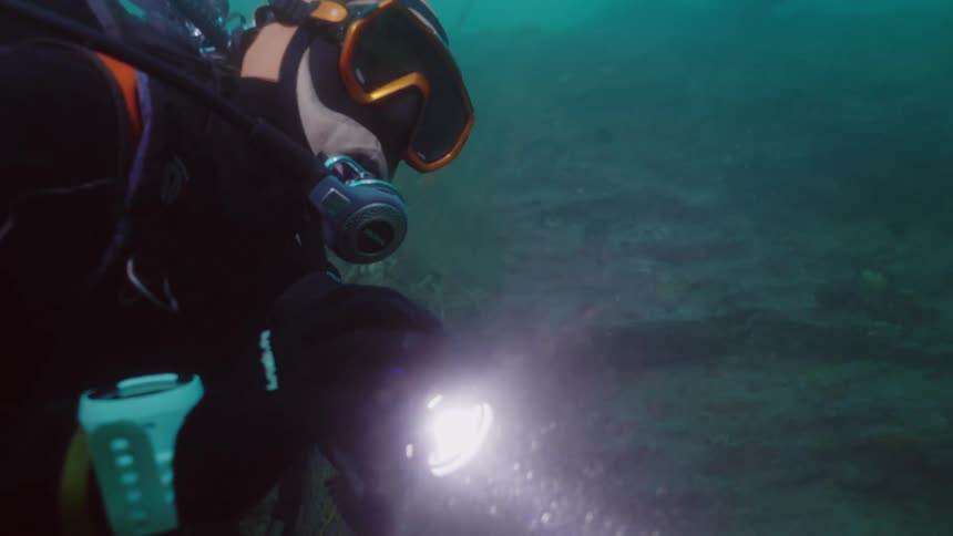 Scuba diver holding flashlight swims through murky waterwhile examining shipwreck