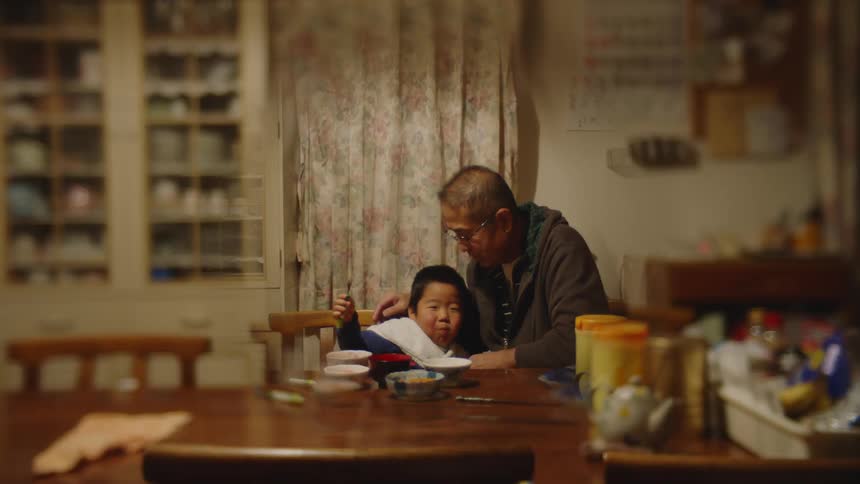 Grandson sitting at table and eat food next to grandfather with glasses and brown jacket