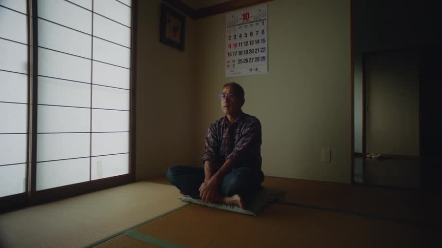 Man with glasses shirt and jeans sits cross legged in room