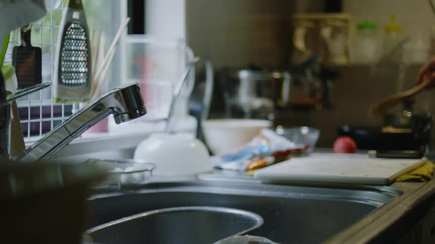 Water is dripping in the kitchen sink as someone is stirring food in a pan cooking on the stove