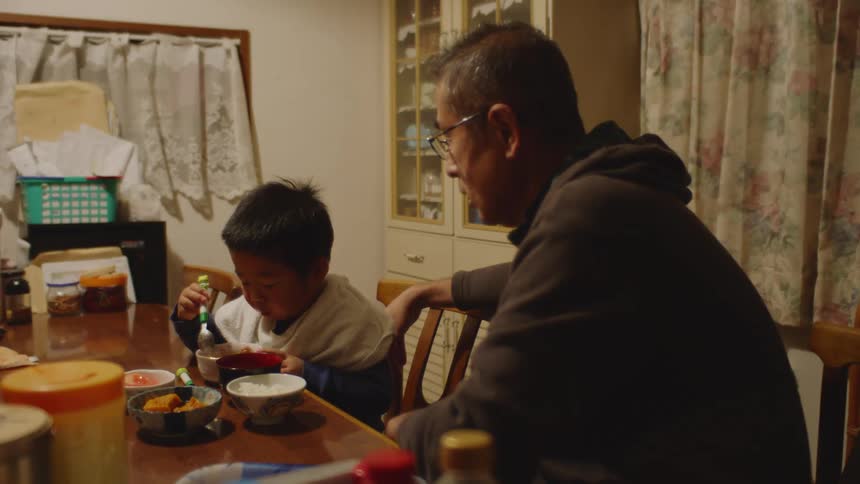 A man sits at a table at home with his son and watches him eat as he talks to him