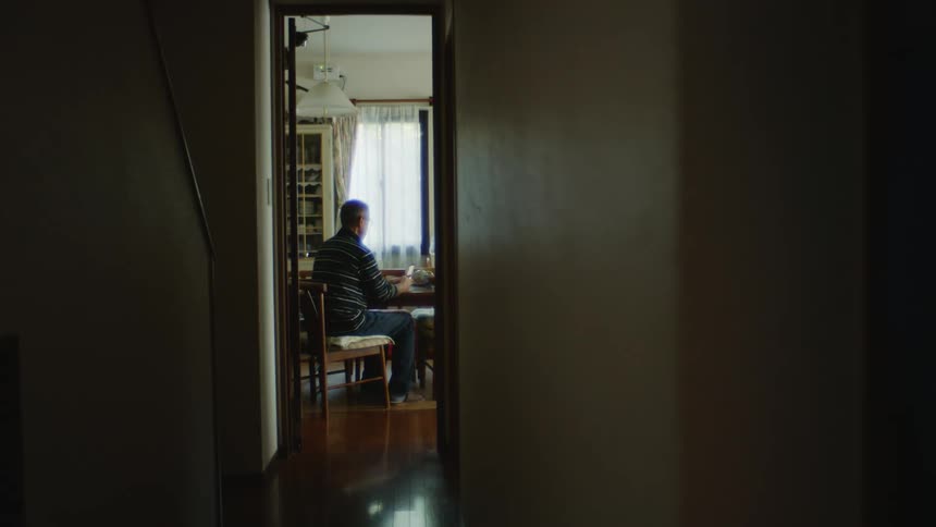 Looking down a hallway a man is sitting at a kitchen table holding his cell phone